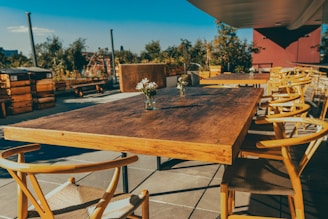 A wooden outdoor dining table surrounded by matching wooden chairs is set on a patio. The setting includes small vases with flowers on the table, under a clear blue sky. The area is surrounded by potted plants and trees, giving a natural and cozy atmosphere.