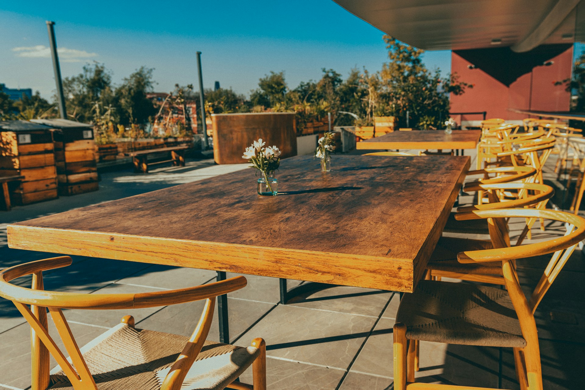 A wooden outdoor dining table surrounded by matching wooden chairs is set on a patio. The setting includes small vases with flowers on the table, under a clear blue sky. The area is surrounded by potted plants and trees, giving a natural and cozy atmosphere.