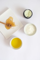 A honey dipper with honey sits on a white square plate. Next to it, there are small bowls containing yellow oil and a white creamy substance. An open jar of greenish cream is also visible.