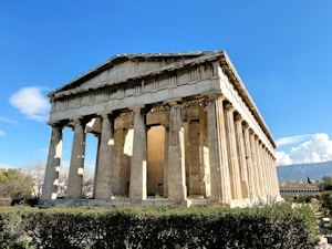 A well-preserved ancient Greek temple with Doric columns surrounded by lush greenery under a clear blue sky. The structure is made of stone, displaying classical architectural design.