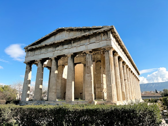 A well-preserved ancient Greek temple with Doric columns surrounded by lush greenery under a clear blue sky. The structure is made of stone, displaying classical architectural design.
