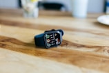 A smartwatch with a black strap is placed on a wooden table. The screen displays various fitness tracking metrics, including step count and activity rings. The background is slightly blurred, showing a white cup and a bouquet of flowers.