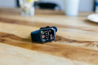 A smartwatch with a black strap is placed on a wooden table. The screen displays various fitness tracking metrics, including step count and activity rings. The background is slightly blurred, showing a white cup and a bouquet of flowers.