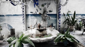 An artistic outdoor setting features a sculpture of a mermaid surrounded by a small water feature. The area is decorated with hanging metallic ornaments and pots placed on the fountain's edge. Potted plants frame the scene, with a painted ocean scene on the wall in the background.