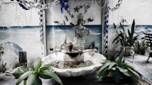 An artistic outdoor setting features a sculpture of a mermaid surrounded by a small water feature. The area is decorated with hanging metallic ornaments and pots placed on the fountain's edge. Potted plants frame the scene, with a painted ocean scene on the wall in the background.