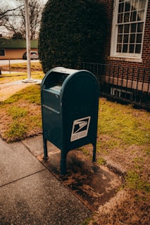 A blue United States Postal Service mailbox stands on a sidewalk near a brick building. The area around the mailbox is covered with grass and patches of dirt. A window with white frames is visible on the building, and a large bush is positioned nearby.