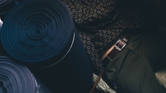 Rolled fabric bolts are arranged, showcasing a rich dark color with a patterned textile in view. A person in dark pants with a detailed belt is adjacent to the fabric.