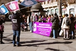 A group of people gathered outdoors, some holding large signs with text. A prominent purple banner in the foreground contains multiple languages, with the main message relating to anti-violence against women. Several individuals are wearing jackets, suggesting cooler weather. A videographer is capturing the event, and a mix of genders and ages is present.