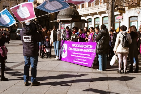 A group of people gathered outdoors, some holding large signs with text. A prominent purple banner in the foreground contains multiple languages, with the main message relating to anti-violence against women. Several individuals are wearing jackets, suggesting cooler weather. A videographer is capturing the event, and a mix of genders and ages is present.