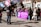 A group of people gathered outdoors, some holding large signs with text. A prominent purple banner in the foreground contains multiple languages, with the main message relating to anti-violence against women. Several individuals are wearing jackets, suggesting cooler weather. A videographer is capturing the event, and a mix of genders and ages is present.
