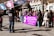 A group of people gathered outdoors, some holding large signs with text. A prominent purple banner in the foreground contains multiple languages, with the main message relating to anti-violence against women. Several individuals are wearing jackets, suggesting cooler weather. A videographer is capturing the event, and a mix of genders and ages is present.