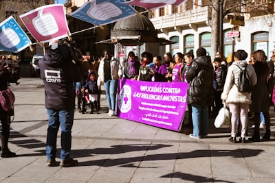 A group of people gathered outdoors, some holding large signs with text. A prominent purple banner in the foreground contains multiple languages, with the main message relating to anti-violence against women. Several individuals are wearing jackets, suggesting cooler weather. A videographer is capturing the event, and a mix of genders and ages is present.