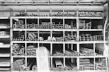 A storage rack filled with various metal pipes and profiles organized by type and size. The rack is divided into compartments, each containing a specific shape or dimension of metal pipe, such as circular, rectangular, and L-shaped sections. The black and white color palette emphasizes the industrial and structured nature of the space.