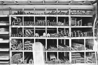 A storage rack filled with various metal pipes and profiles organized by type and size. The rack is divided into compartments, each containing a specific shape or dimension of metal pipe, such as circular, rectangular, and L-shaped sections. The black and white color palette emphasizes the industrial and structured nature of the space.