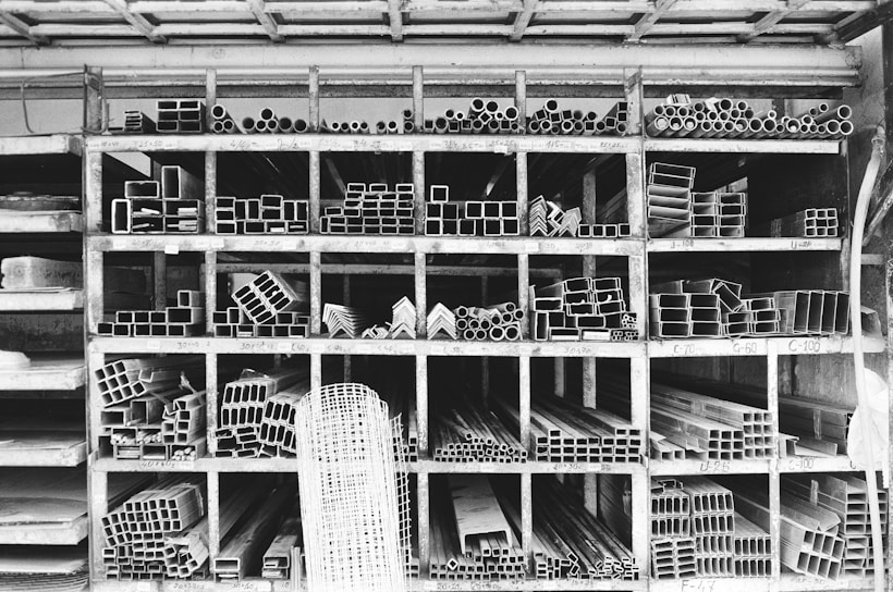 A storage rack filled with various metal pipes and profiles organized by type and size. The rack is divided into compartments, each containing a specific shape or dimension of metal pipe, such as circular, rectangular, and L-shaped sections. The black and white color palette emphasizes the industrial and structured nature of the space.