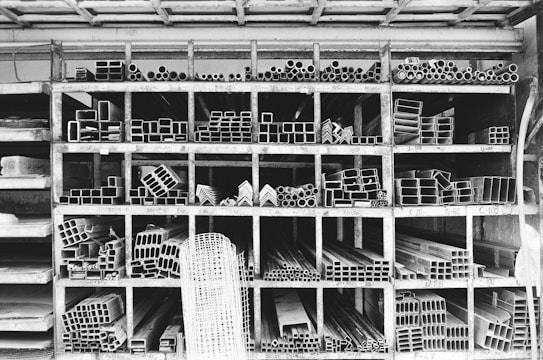A storage rack filled with various metal pipes and profiles organized by type and size. The rack is divided into compartments, each containing a specific shape or dimension of metal pipe, such as circular, rectangular, and L-shaped sections. The black and white color palette emphasizes the industrial and structured nature of the space.