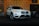A white SUV is displayed in a showroom with a patterned backdrop featuring the letters 'GA'. The vehicle is highlighted, showcasing its front grille and headlights, while another vehicle is partially visible in the background.