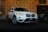A white SUV is displayed in a showroom with a patterned backdrop featuring the letters 'GA'. The vehicle is highlighted, showcasing its front grille and headlights, while another vehicle is partially visible in the background.
