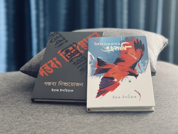 Two books are placed on a textured gray surface. The book on the left has a black cover with bold red and black text in an artistic font. The book on the right features a white cover with an illustration of a large red and black bird in flight, with additional text in a stylized script. The background includes a soft-focus curtain, creating a calm ambiance.