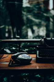 A vintage facsimile machine resting on a wooden desk beside old newspapers and a rotary phone, evoking the 1980s era.