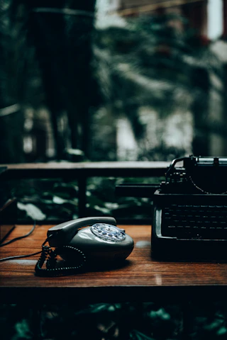 A vintage facsimile machine resting on a wooden desk beside old newspapers and a rotary phone, evoking the 1980s era.