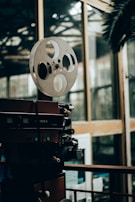 A vintage film projector with large film reels sits in a dimly lit room, surrounded by reflective glass windows. The setting appears to be an indoor space with a mixture of modern and retro elements.
