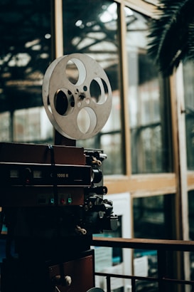 A vintage film projector with large film reels sits in a dimly lit room, surrounded by reflective glass windows. The setting appears to be an indoor space with a mixture of modern and retro elements.
