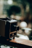 A vintage film projector with a textured, dark surface and a metallic lens protruding from the front. It is placed on a wooden surface with a blurred green and brown background that suggests an outdoor setting.