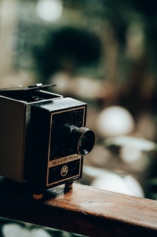 A vintage film projector with a textured, dark surface and a metallic lens protruding from the front. It is placed on a wooden surface with a blurred green and brown background that suggests an outdoor setting.
