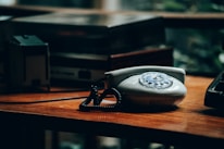 A vintage rotary dial telephone sits on a wooden desk, surrounded by other retro electronic equipment like an old amplifier or cassette deck. The lighting is dim, giving the scene an atmospheric and nostalgic feel.