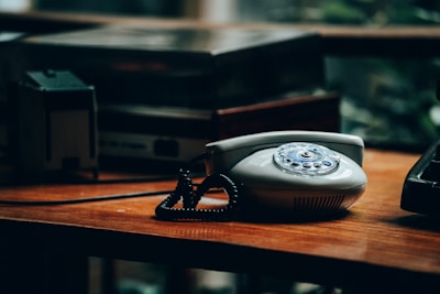 A vintage rotary dial telephone sits on a wooden desk, surrounded by other retro electronic equipment like an old amplifier or cassette deck. The lighting is dim, giving the scene an atmospheric and nostalgic feel.