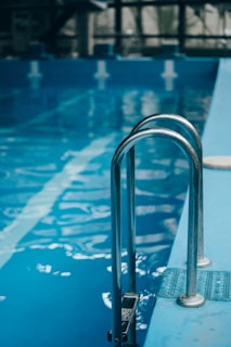 Metal pool ladder leading into clear blue water of an indoor swimming pool. The water reflects soft natural light, with visible lane markers on the pool floor.