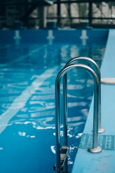 Metal pool ladder leading into clear blue water of an indoor swimming pool. The water reflects soft natural light, with visible lane markers on the pool floor.