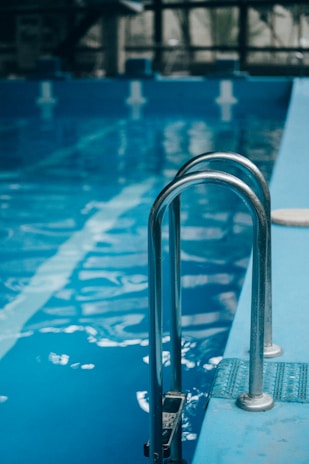 Metal pool ladder leading into clear blue water of an indoor swimming pool. The water reflects soft natural light, with visible lane markers on the pool floor.