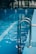Metal pool ladder leading into clear blue water of an indoor swimming pool. The water reflects soft natural light, with visible lane markers on the pool floor.