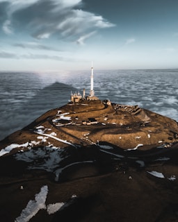 A mountainous landscape rising above a sea of clouds, featuring a large antenna tower on top of the hill. The terrain is dotted with patches of snow, and winding roads lead up to the structure. The sky above is dramatic with scattered clouds.