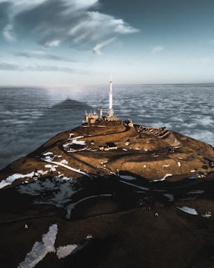 A mountainous landscape rising above a sea of clouds, featuring a large antenna tower on top of the hill. The terrain is dotted with patches of snow, and winding roads lead up to the structure. The sky above is dramatic with scattered clouds.