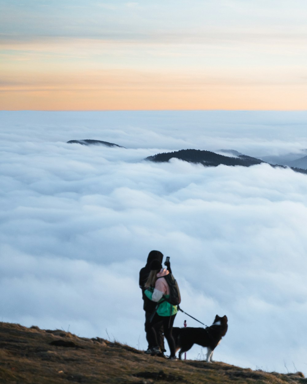 um homem e uma mulher com um cão em uma colina acima das nuvens