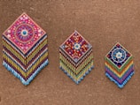 Three intricately beaded wall hangings with diamond shapes are displayed against a textured brown background. Each piece features colorful geometric and floral patterns, with vibrant strands of beads hanging below. The designs include detailed flower motifs and symmetrical arrangements, showcasing craftsmanship and artistry.