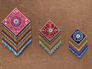 Three intricately beaded wall hangings with diamond shapes are displayed against a textured brown background. Each piece features colorful geometric and floral patterns, with vibrant strands of beads hanging below. The designs include detailed flower motifs and symmetrical arrangements, showcasing craftsmanship and artistry.