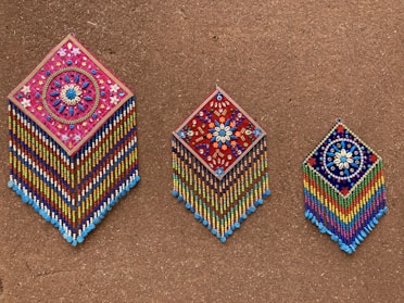 Three intricately beaded wall hangings with diamond shapes are displayed against a textured brown background. Each piece features colorful geometric and floral patterns, with vibrant strands of beads hanging below. The designs include detailed flower motifs and symmetrical arrangements, showcasing craftsmanship and artistry.
