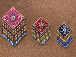 Three intricately beaded wall hangings with diamond shapes are displayed against a textured brown background. Each piece features colorful geometric and floral patterns, with vibrant strands of beads hanging below. The designs include detailed flower motifs and symmetrical arrangements, showcasing craftsmanship and artistry.