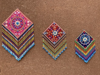 Three intricately beaded wall hangings with diamond shapes are displayed against a textured brown background. Each piece features colorful geometric and floral patterns, with vibrant strands of beads hanging below. The designs include detailed flower motifs and symmetrical arrangements, showcasing craftsmanship and artistry.