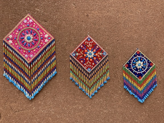 Three intricately beaded wall hangings with diamond shapes are displayed against a textured brown background. Each piece features colorful geometric and floral patterns, with vibrant strands of beads hanging below. The designs include detailed flower motifs and symmetrical arrangements, showcasing craftsmanship and artistry.