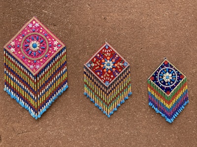 Three intricately beaded wall hangings with diamond shapes are displayed against a textured brown background. Each piece features colorful geometric and floral patterns, with vibrant strands of beads hanging below. The designs include detailed flower motifs and symmetrical arrangements, showcasing craftsmanship and artistry.
