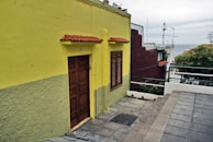 A vibrant yellow house with a wooden door and window shutters, featuring a small overhang with red tiles. The house is bordered by a gray sidewalk leading toward a view of the ocean in the distance. On the right, neighboring buildings include a dark red structure, roof antennas, greenery, and a metal railing.