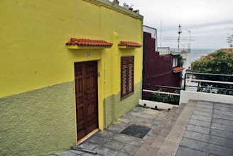 A vibrant yellow house with a wooden door and window shutters, featuring a small overhang with red tiles. The house is bordered by a gray sidewalk leading toward a view of the ocean in the distance. On the right, neighboring buildings include a dark red structure, roof antennas, greenery, and a metal railing.