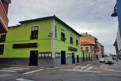 A vibrant lime-green building stands at the corner of a street, housing a restaurant. The building features large windows with flowerpots on the sills and a dark roof. A street sign is visible next to a pedestrian crossing, and several buildings line the street in the background.