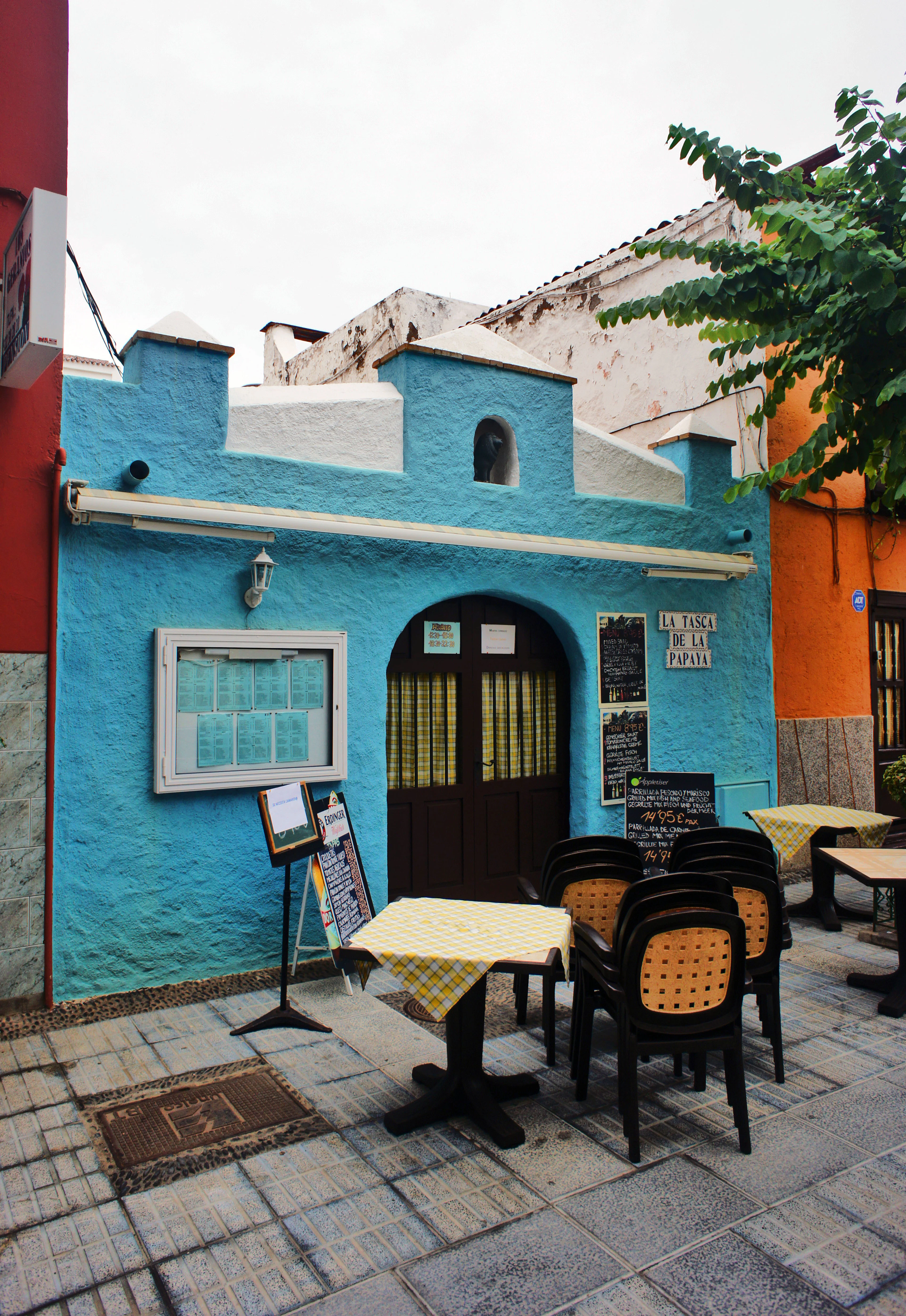 A small sky-blue tasca in Puerto de la Cruz, Tenerife, chalkboard menus propped either side of its dark wooden door and yellow gingham-covered tables set out on the tiled pavement beside an orange neighbouring wall.