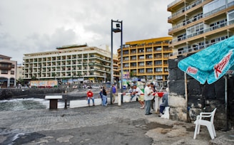 A coastal urban area with several multistory buildings, one of which features yellow balconies. People are gathered around on a cobblestone area near the sea. An advertisement canopy is visible along with a white plastic chair. The scene appears lively but the sky is overcast.
